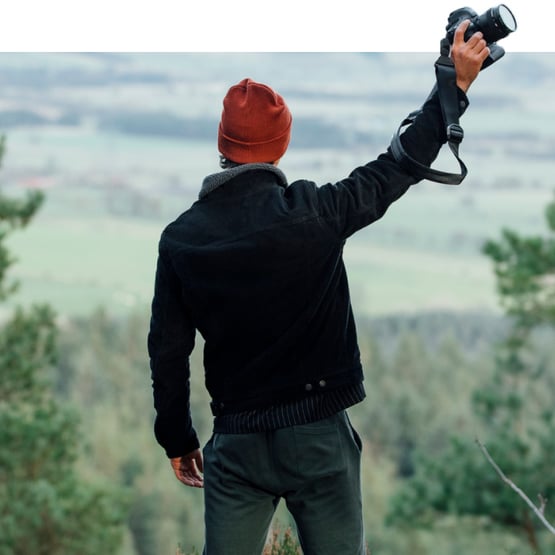 Hero image of a man at the top of a forest hill holding a camera in the air