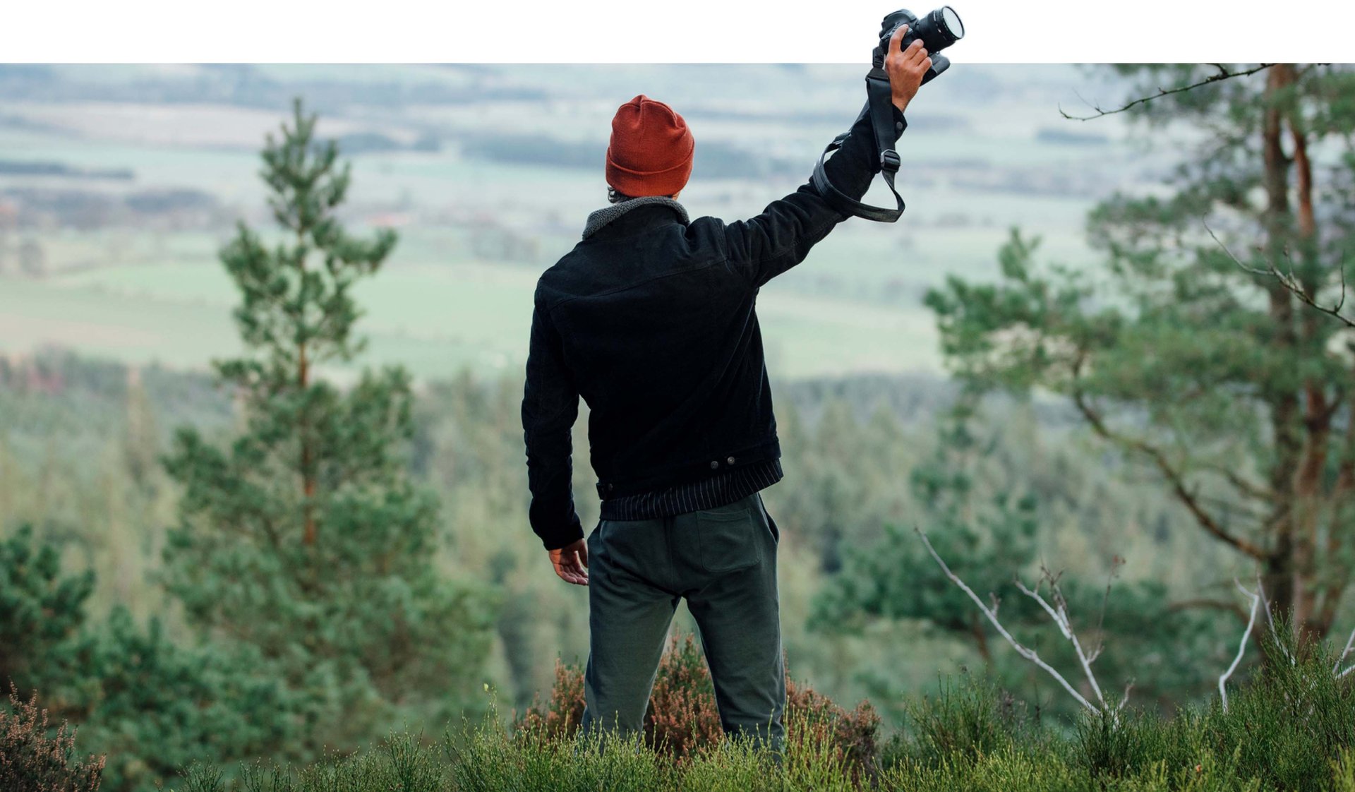 Hero image of a man at the top of a forest hill holding a camera in the air